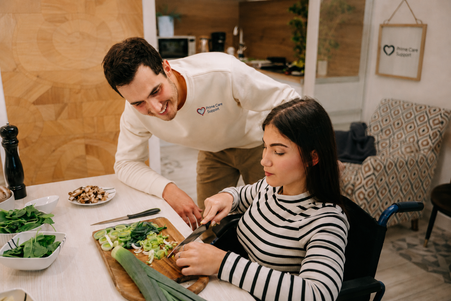 A caregiver and a woman who uses a wheelchair prepare a meal together in a sunlit kitchen, smiling and chatting.